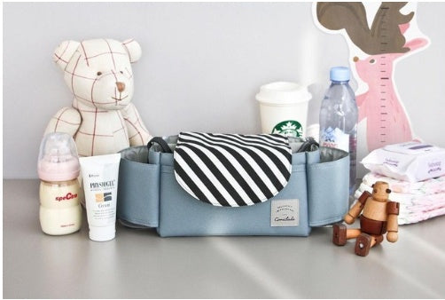 Stroller organiser bag styled on a table with baby bottle, cream, teddy bear, wipes, and wooden toy